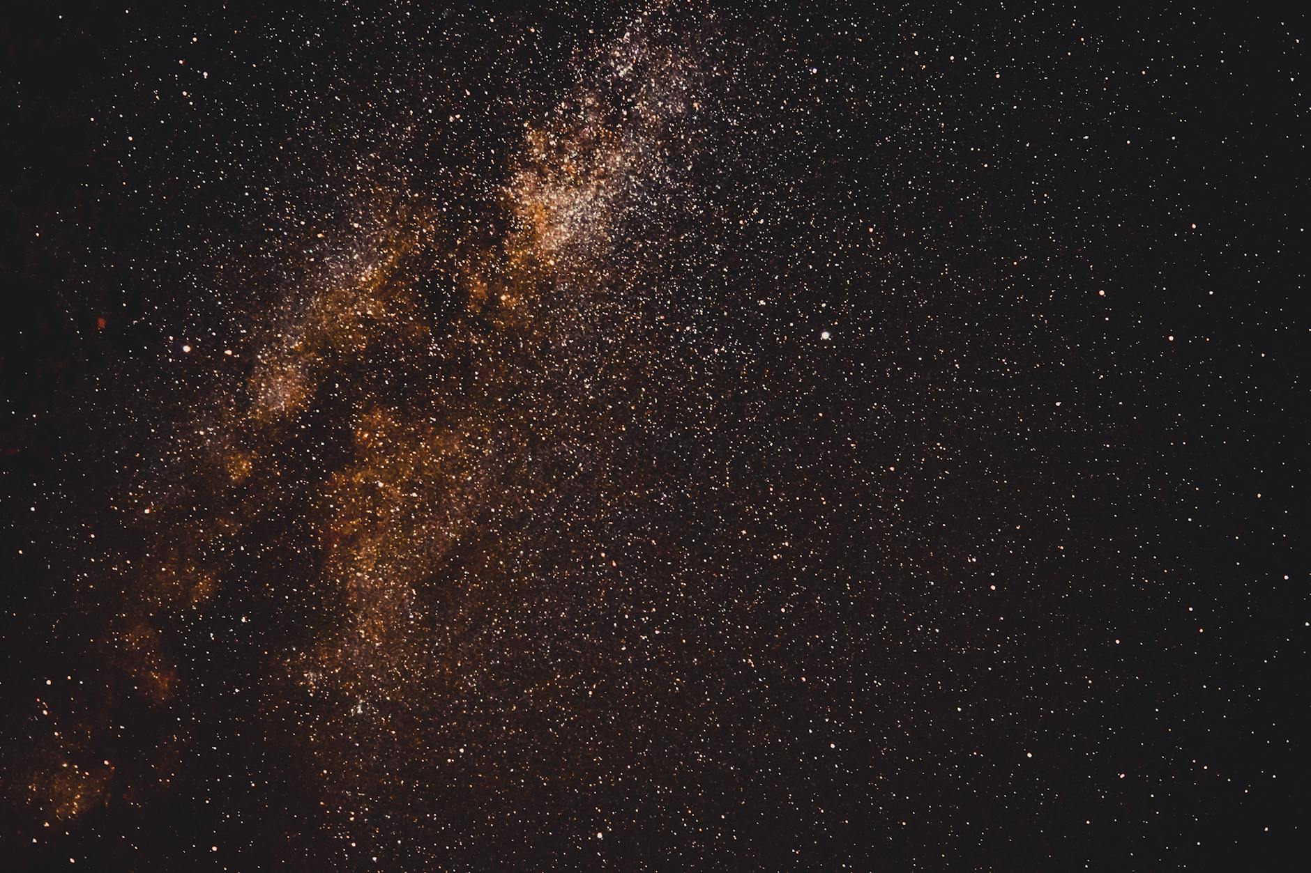 A photo of space taken by Delcho Dichev on Pexels.com. It is a piece of astrophotography that depicts a galaxy on a dark background. The space looks largely black with some light orange and rust coloured lighting and clouds. There are many bright white sparks pictured.